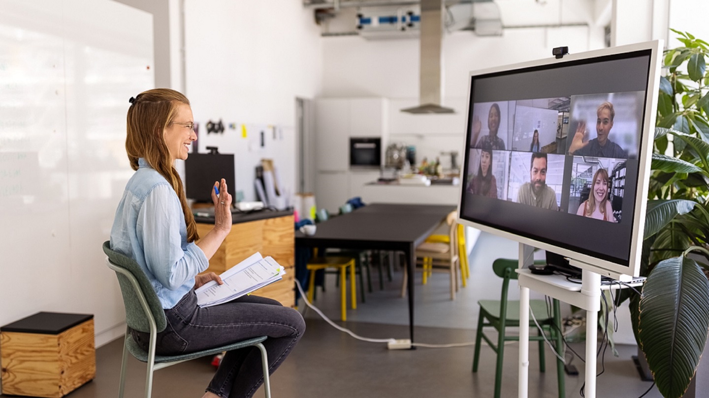 Female professional having online meeting with team seen on big television screen. Businesswoman in hybrid office space having web conference with coworkers.
