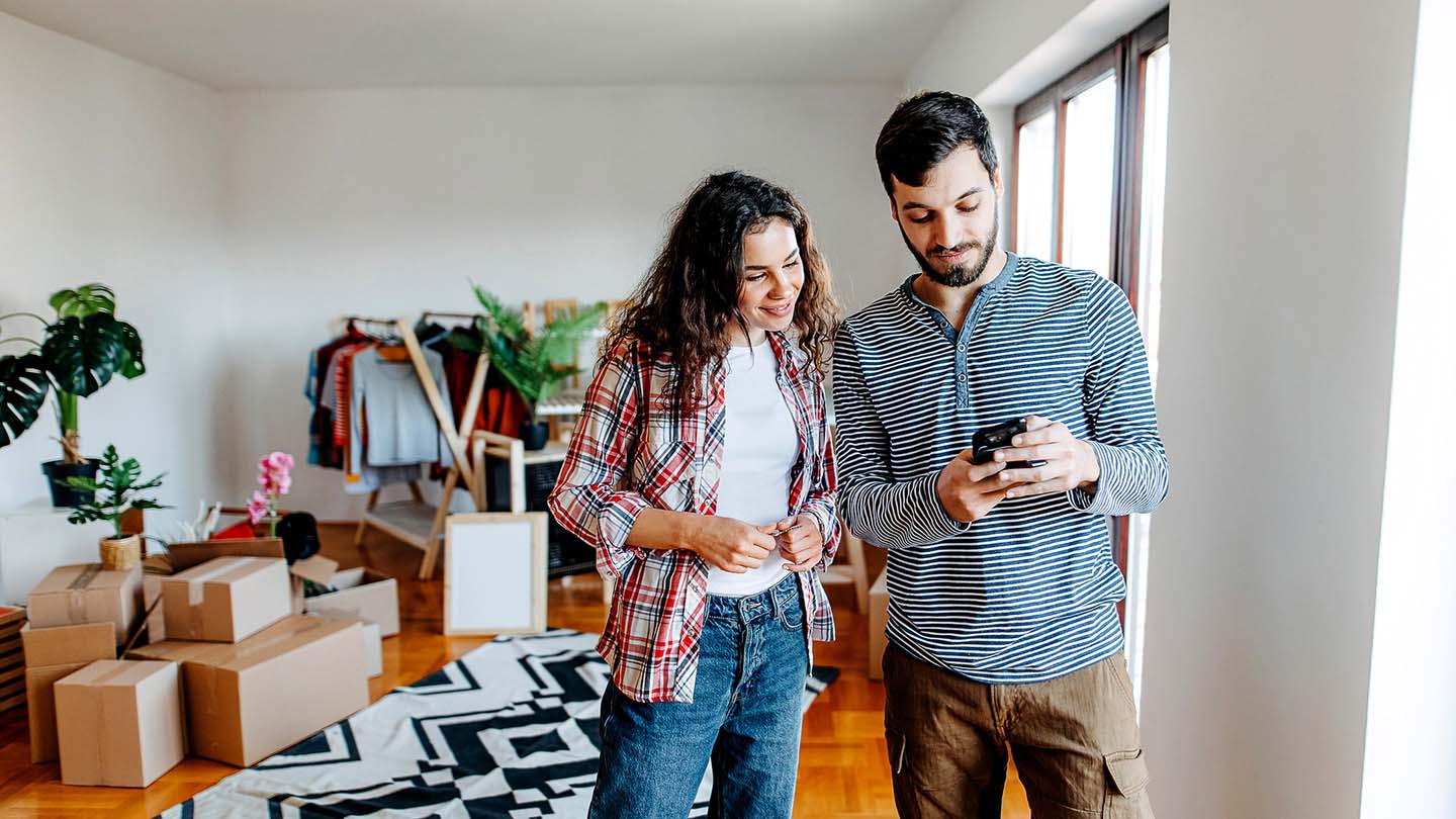 A man and woman look at rental listings on a phone in their apartment