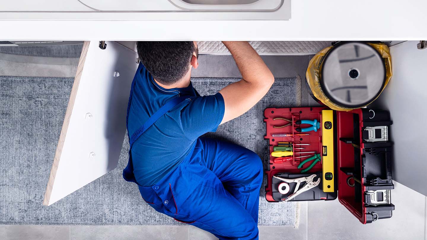 A man works on a repair in a cabinet beneath a kitchen sink.
