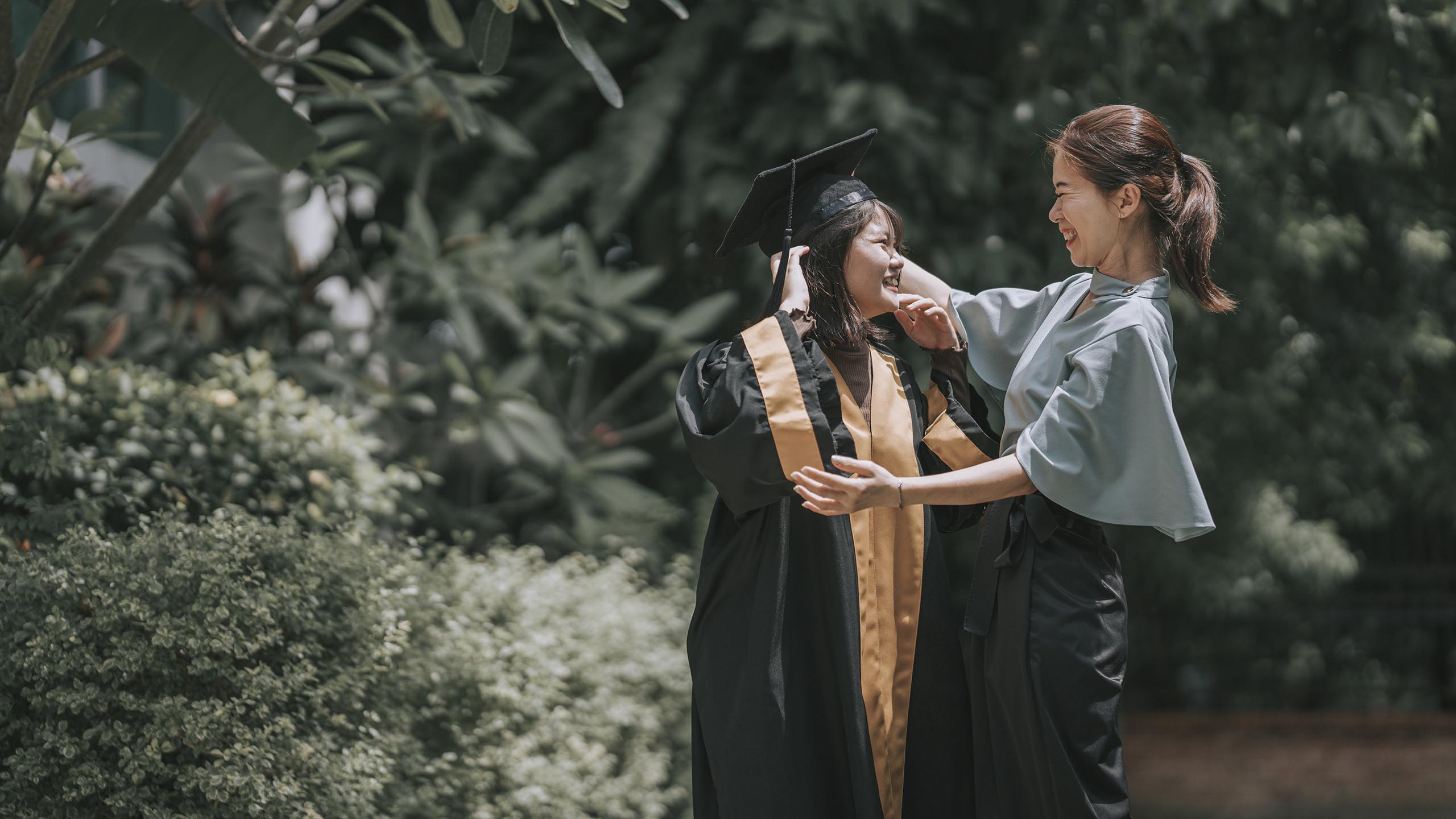 Proud Asian Chinese Mother adjusting mortarboard for daughter in college graduation gown outside college buildings getting ready for graduation ceremony