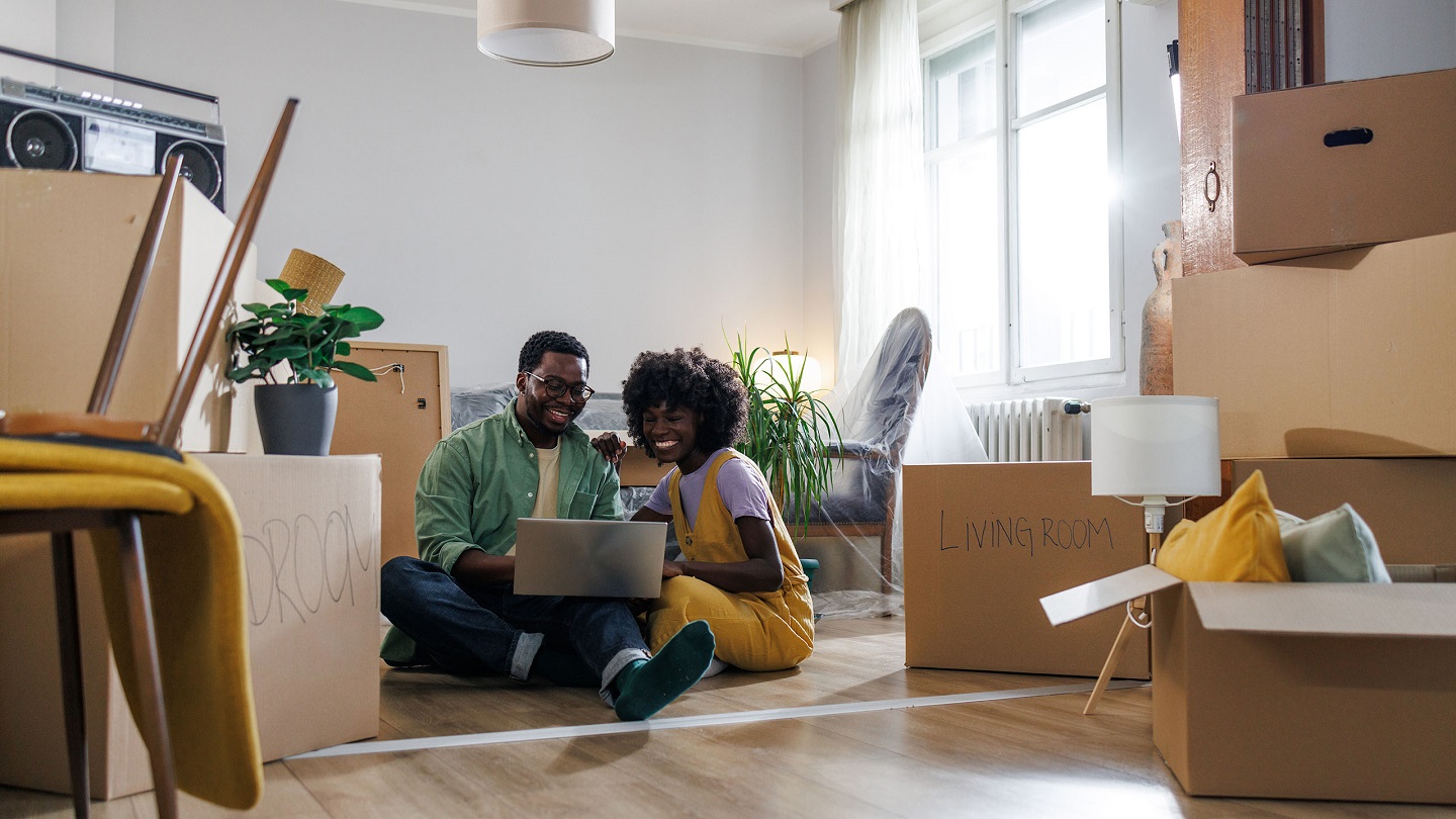 Photo of a cheerful African-American couple relaxing on the floor of their new apartment. They are surfing the net on their laptop.