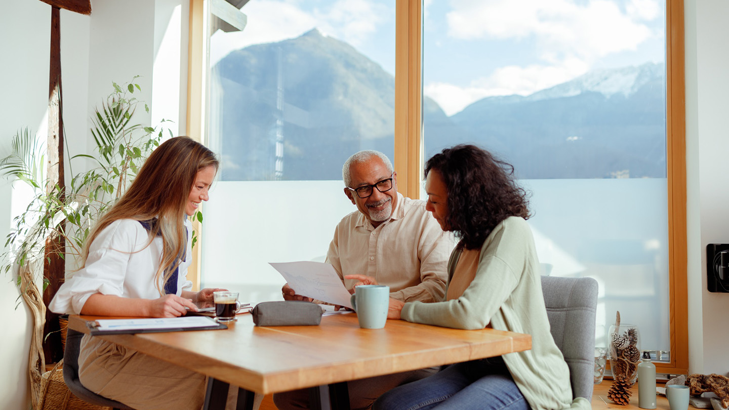 A woman, likely a financial advisor, is seated with a senior couple in a bright room with a stunning mountain view, indicative of a home in Slovenia. They are engaged in conversation, with documents laid out on the table, pointing to a discussion about pension insurance. The advisor is presenting information, while the couple listens intently, considering the options for their retirement planning.