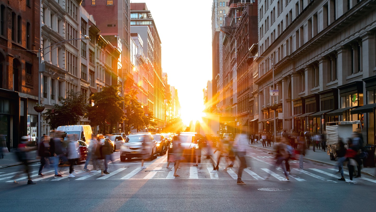 Crowds of busy people walking through the intersection of 5th Avenue and 23rd Street in Manhattan, New York City with bright sunset background