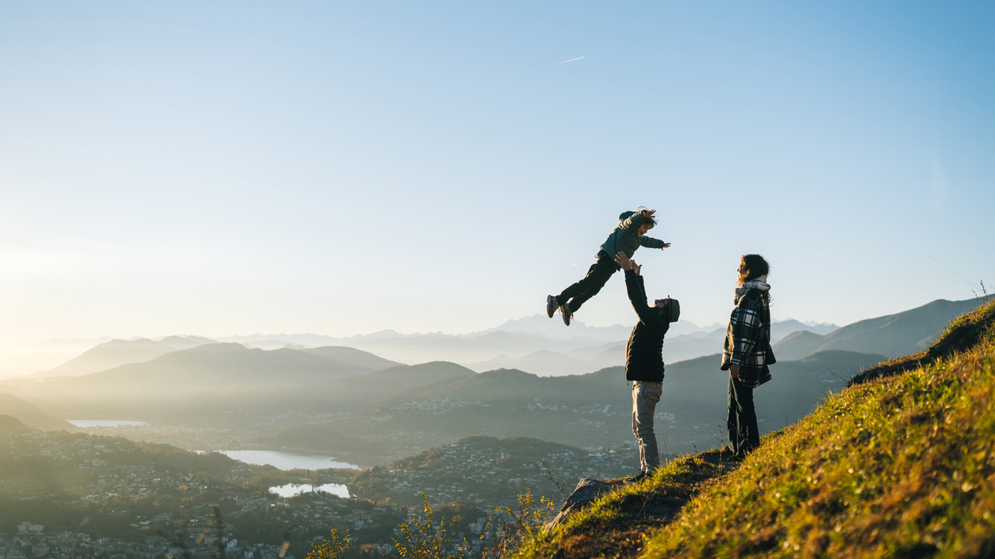 Father throws son in air above head on mountain top at sunset, mother watches